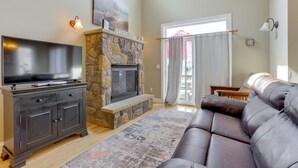 A living room with a stone fireplace, a flat-screen TV, a dark leather sofa, and a large window with curtains. - < Half-Mi to Lake: Townhome w/ Hot Tub in McHenry! (McHenry)