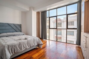 A bedroom with a bed, a large window, and wooden flooring.