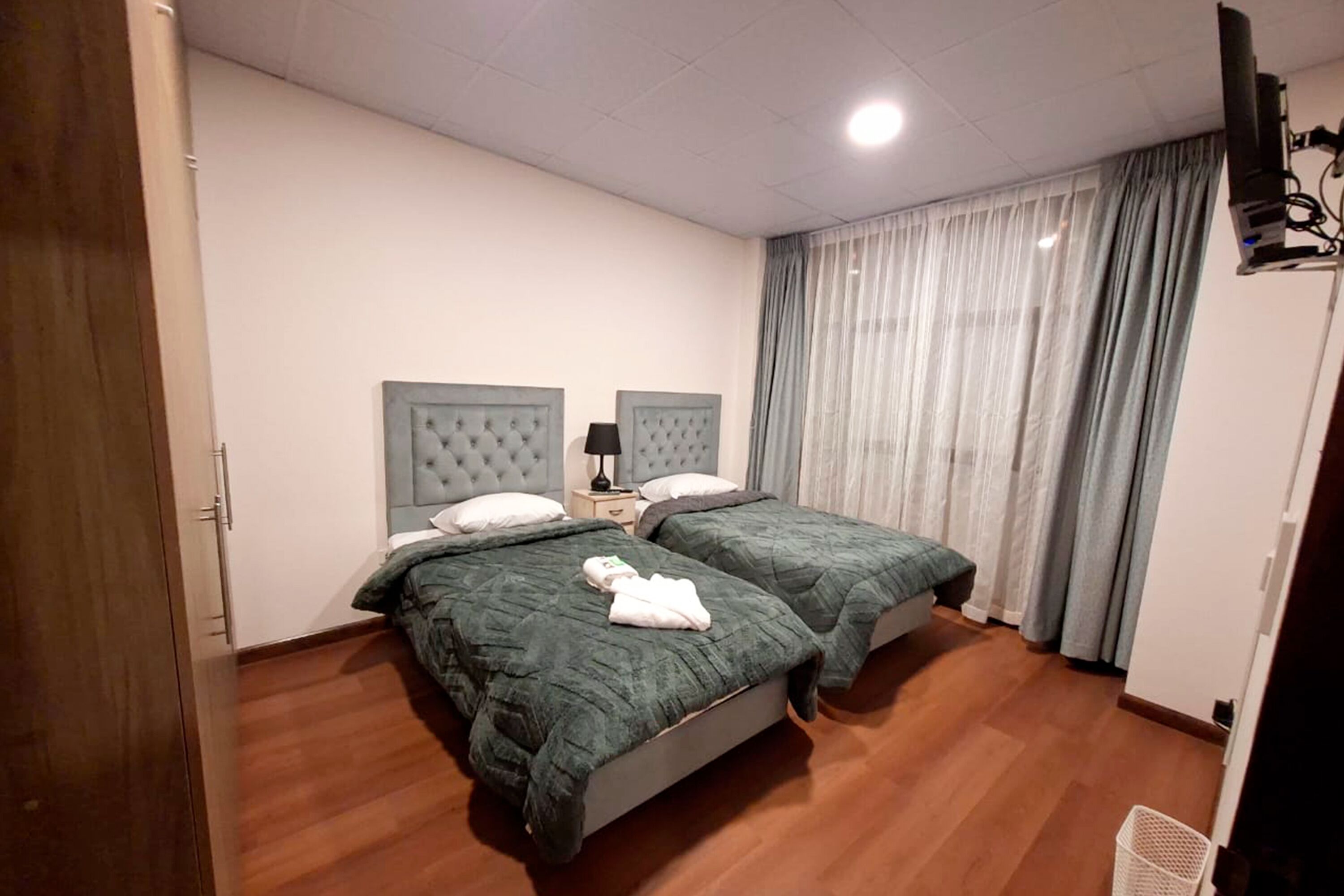 A hotel room with two beds, a wooden floor, a white wall, a ceiling light, and a window with curtains.