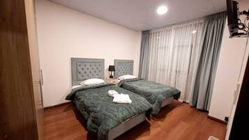 A hotel room with two beds, a wooden floor, a white wall, a ceiling light, and a window with curtains.
