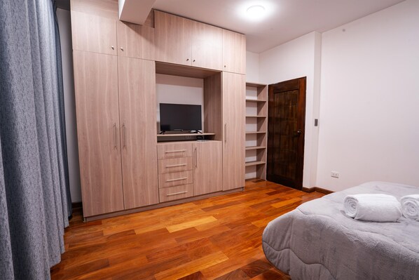 A bedroom with a wooden wardrobe, a bed with a grey duvet, and a television on a shelf.
