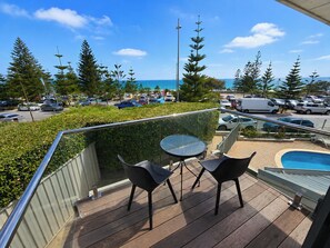 A balcony with a table and chairs, overlooking a parking area and a swimming pool.