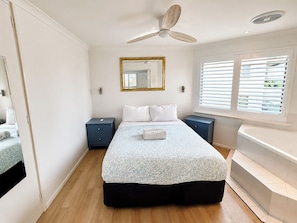 A bedroom with a bed, bedside tables, a ceiling fan, a window with blinds, and a mirror. - Sunset Spa Escape - Ocean Views (Scarborough)