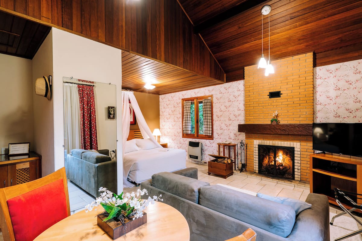 Una habitación acogedora con una cama, un sofá gris y un televisor. Hay una chimenea, un techo de madera y una zona de comedor con una mesa y sillas.