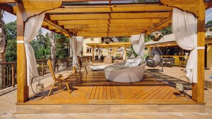 Un espacio de estar al aire libre techado con terraza de madera, sofá redondo de mimbre y una silla colgante.