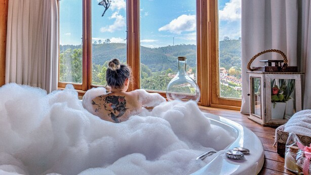 Una habitación con un gran ventanal, una bañera llena de burbujas y vistas a un paisaje boscoso.