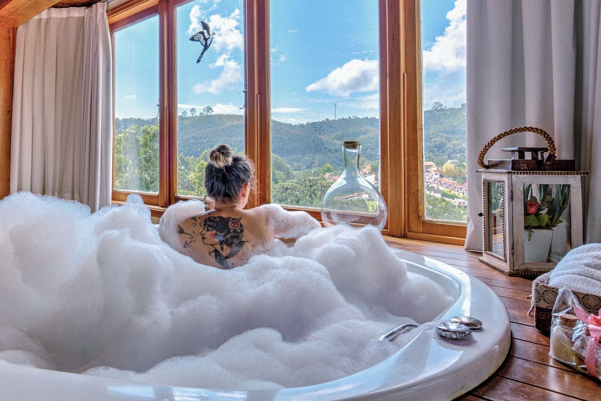 Una habitación con un gran ventanal, una bañera llena de burbujas y vistas a un paisaje boscoso.