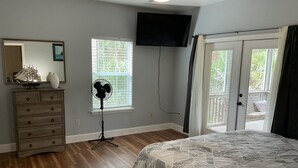 A bedroom with a bed, dresser, fan, TV, and a window with blinds.