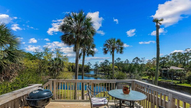 A wooden deck with a grill, chairs, and a table overlooking a lake and palm trees.