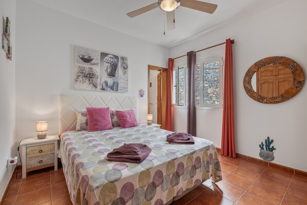 Een slaapkamer met een bed, nachtkastje, decoratieve muurkunst, gordijnen en een plafondventilator.