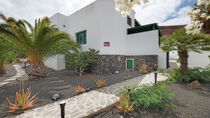 A white building with a stone base, surrounded by tropical plants and a paved pathway.