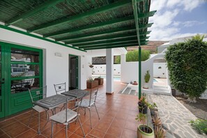 Un patio al aire libre con una mesa y sillas, rodeado de plantas en macetas y una pared blanca.