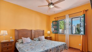 Un dormitorio con cama, mesitas de noche, ventilador de techo y ventana con cortinas.