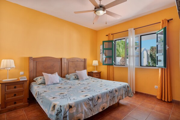 Un dormitorio con cama, mesitas de noche, ventilador de techo y ventana con cortinas.