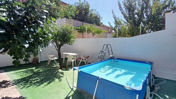 Une piscine avec un escalier, entourée d’un mur blanc et dotée d’un revêtement de sol vert. On y trouve deux chaises blanches et une petite table.