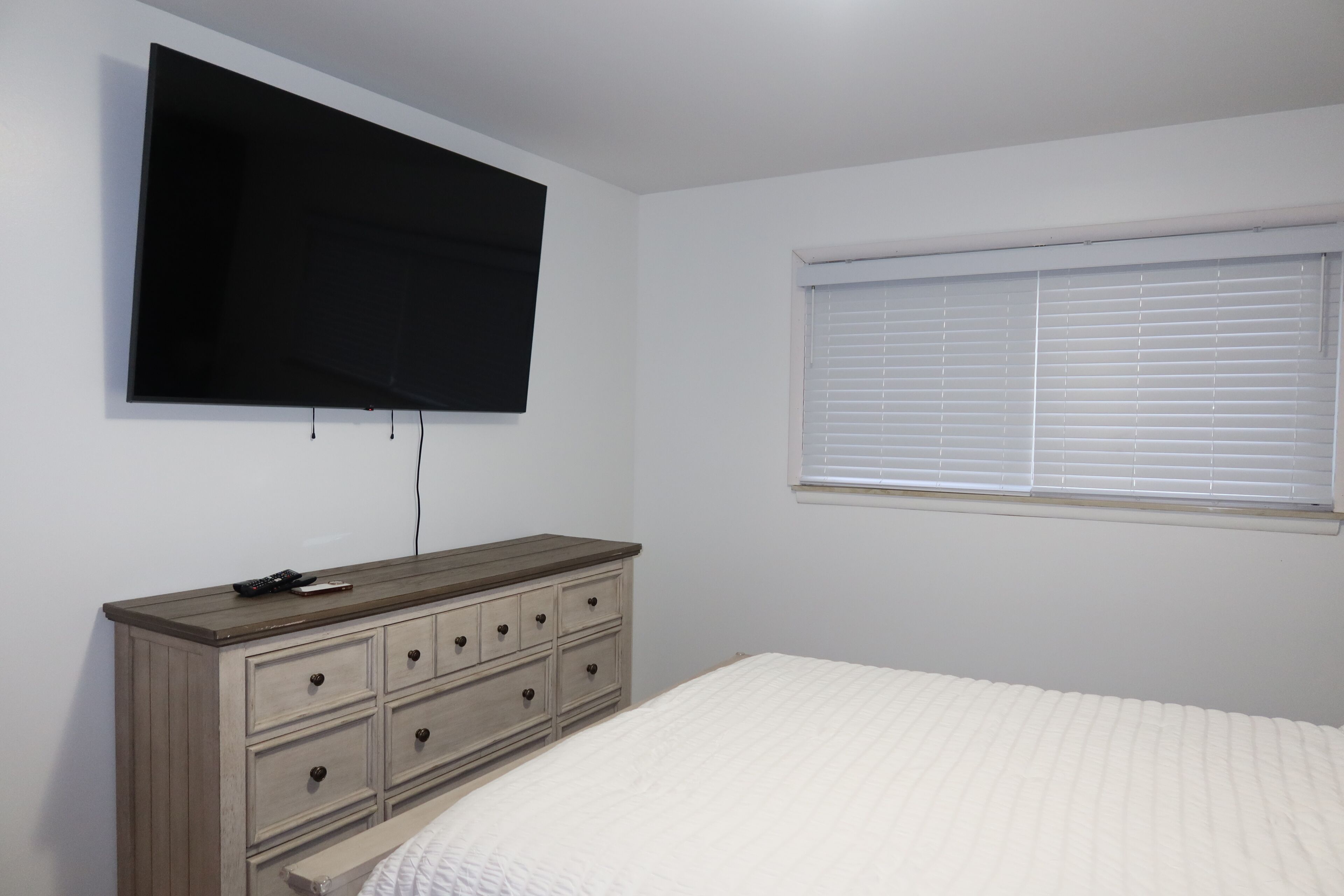 A bedroom with a bed, a dresser, a television mounted on the wall, and a window with blinds.