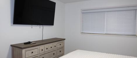 A bedroom with a bed, a dresser, a television mounted on the wall, and a window with blinds.
