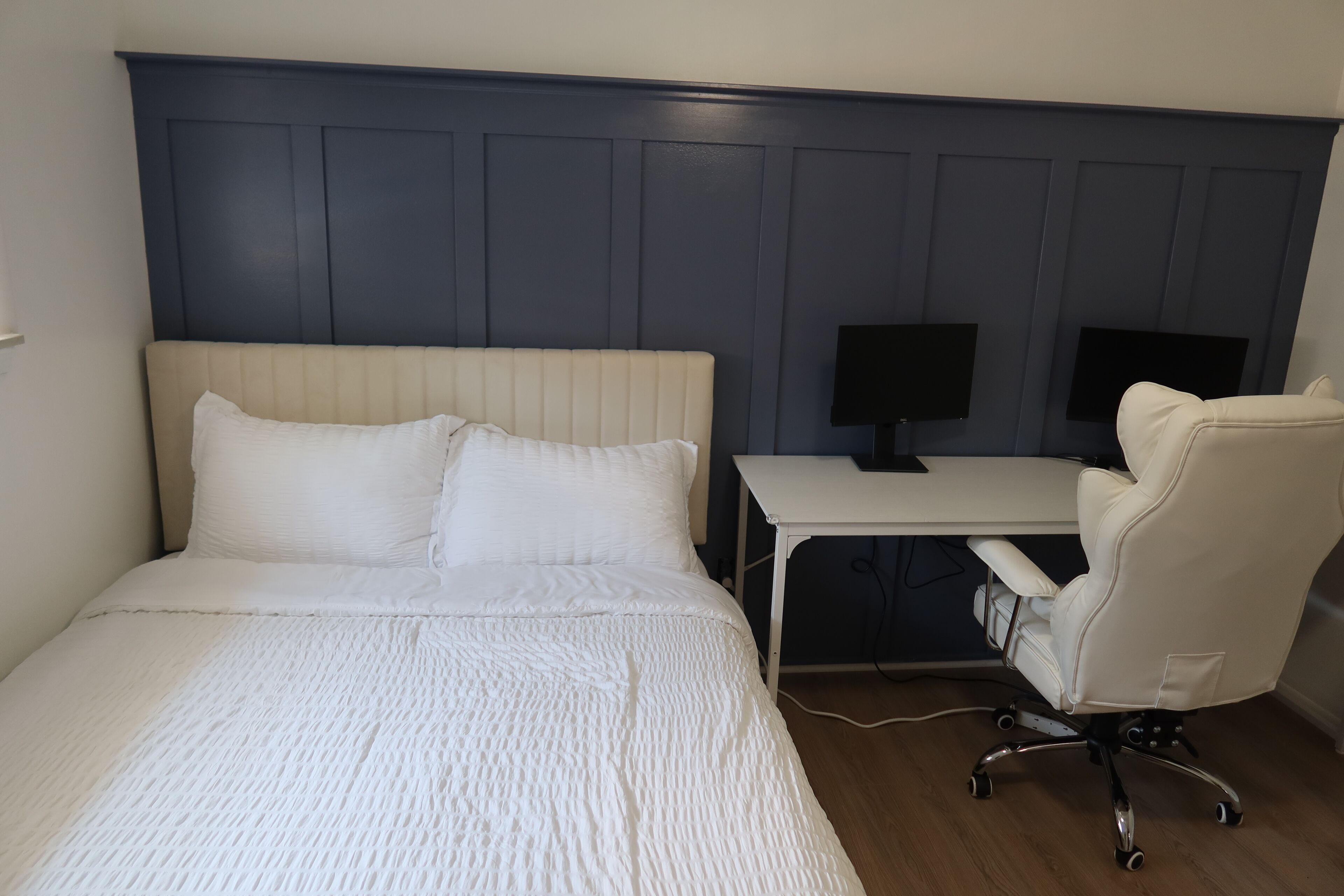 A hotel room with a bed, a desk, a chair, and two monitors.