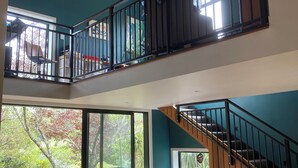 A spacious room with a wooden ceiling, a balcony with a black railing, a dining area with a table and chairs, and a large sliding glass door leading to an outdoor deck.