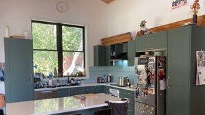 A kitchen with teal cabinets, a central island with bar stools, a refrigerator with magnets, and a window with greenery outside.