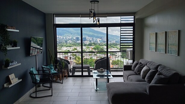 Een moderne woonkamer met een groot raam, een grijze bank, een glazen salontafel en een zwarte fauteuil.