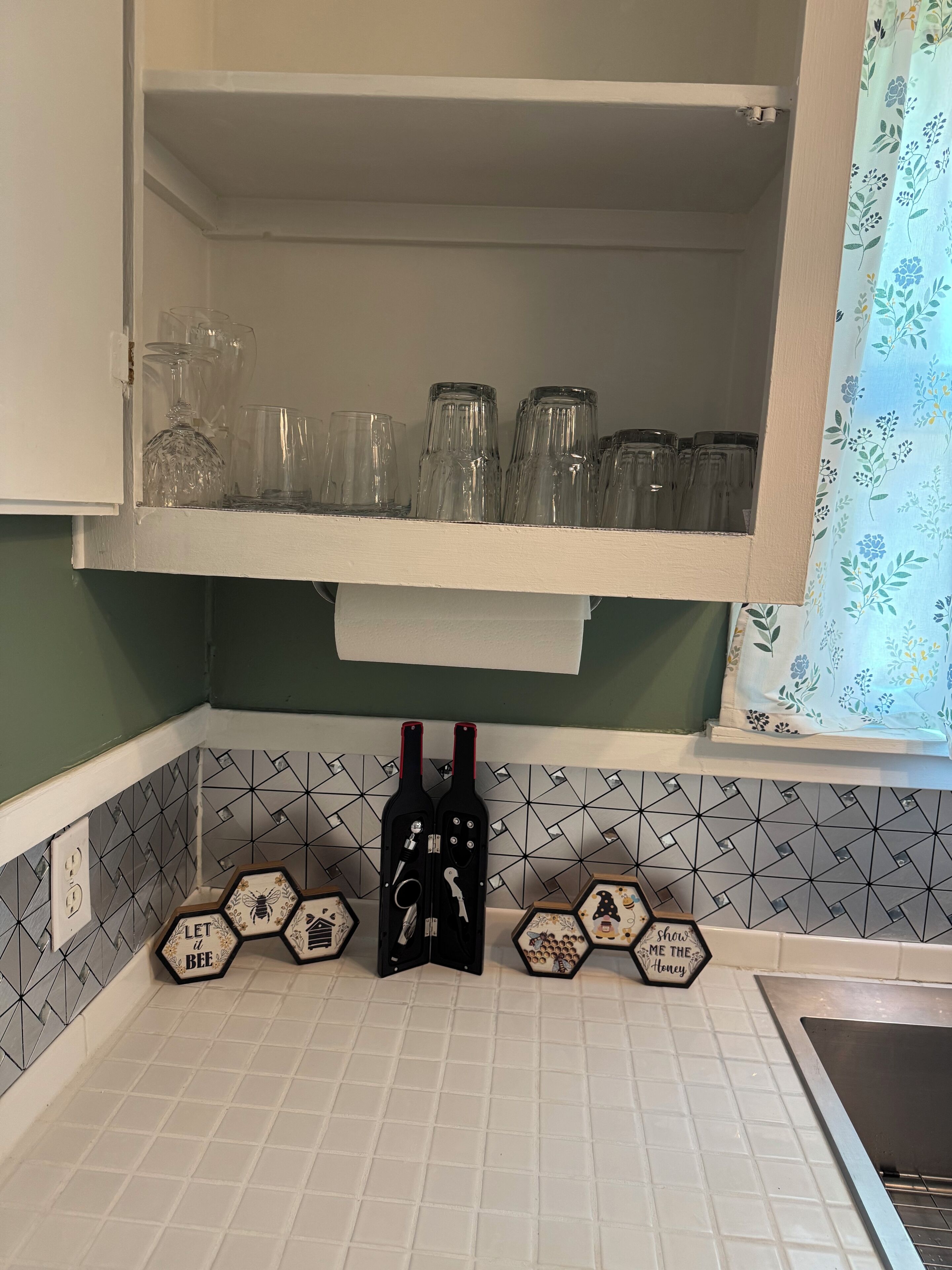A kitchen with a white cabinet shelf holding glassware and a black wine bottle with a corkscrew. Below, hexagonal tiles with bee-themed decorations are visible.