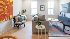 A modern living room with a sofa, a coffee table, and a TV. There are two windows and a ceiling fan.