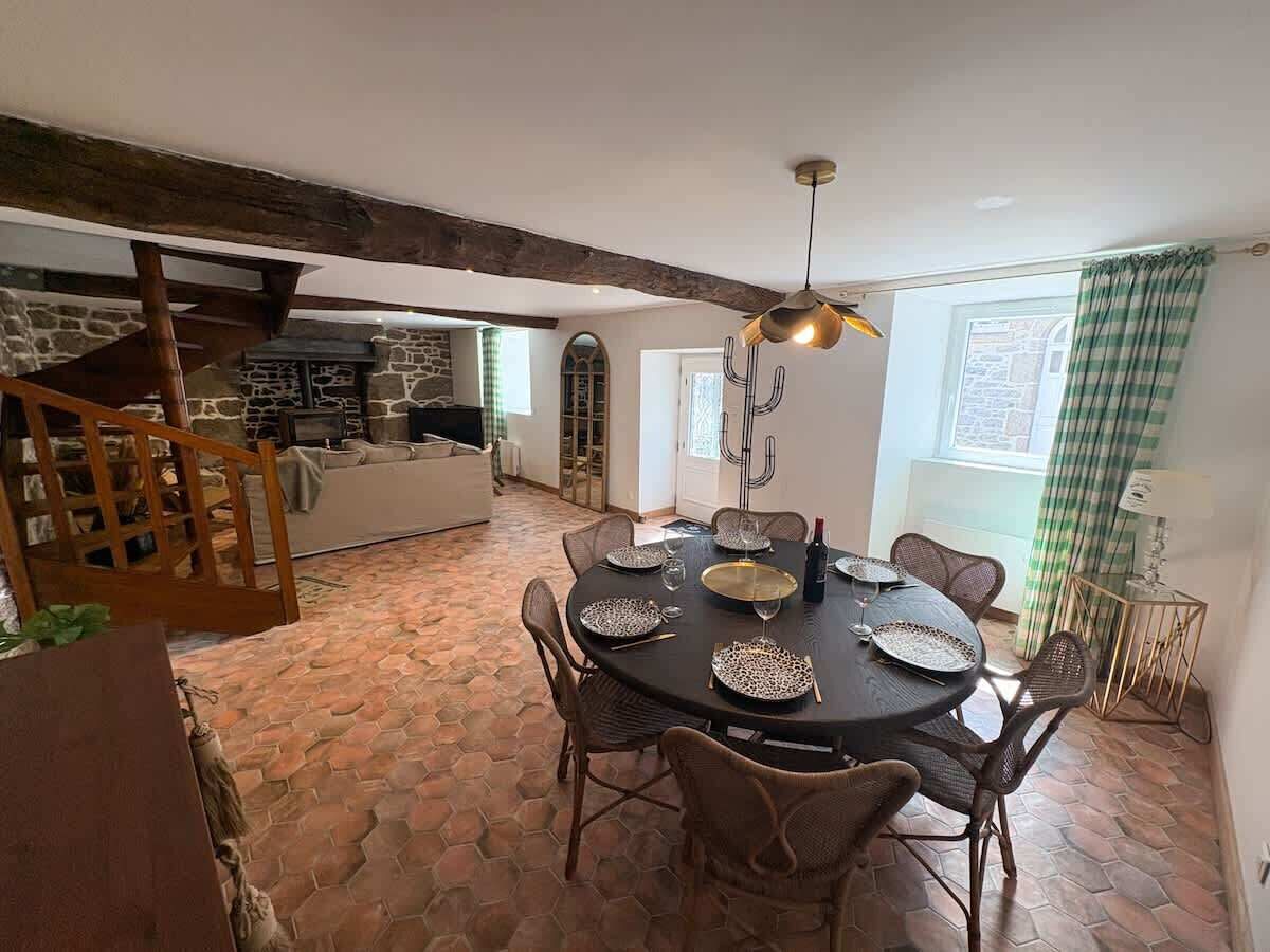 A dining area with a round table set for four, surrounded by wicker chairs. A chandelier hangs above the table. The room features exposed wooden beams and a stone wall in the background.