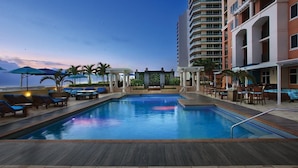 Zona de piscina con sillones de descanso y sombrillas, rodeada por una terraza de madera y un edificio con balcones.