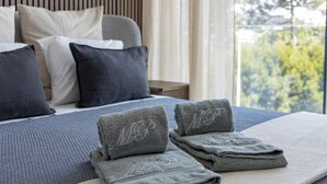 Una cama bien hecha con ropa de cama y almohadas grises, una mesita de noche y vistas al exterior con vegetación.