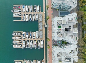 An aerial view of a marina with numerous boats docked at wooden piers. Adjacent to the marina is a modern residential building with a unique architectural design, featuring multiple balconies and terraces.