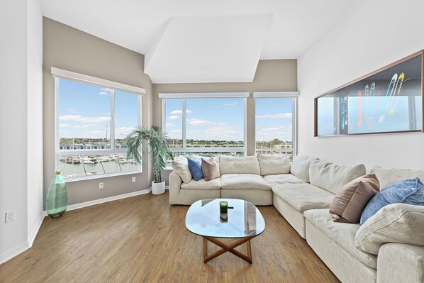 A spacious living room with a large window, a beige sofa, a glass coffee table, and a potted plant.