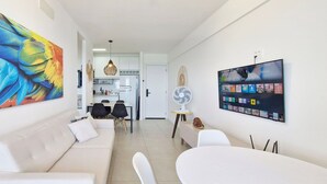 A modern living room with a white sofa, a dining area with black chairs, a large colorful painting, a wall-mounted TV, and a fan.