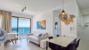 A modern living room with a sofa, dining table, and chairs, a large window with a view of the sea, and a kitchen area with a washing machine.