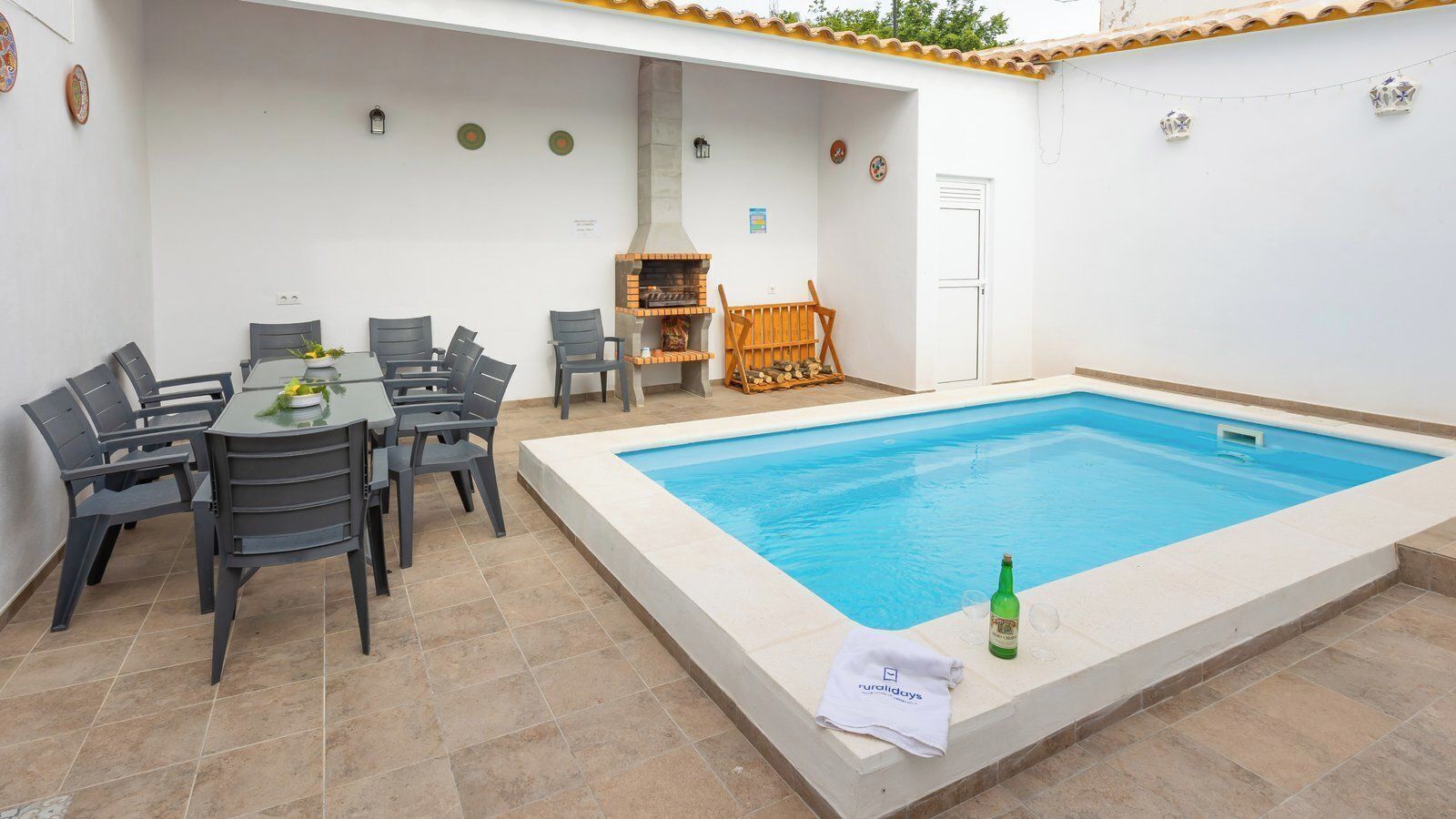 Zona de piscina con mesa y sillas, una parrilla y suelo de baldosas.