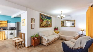 Una sala de estar con un sofá, sillas, una mesita con una planta, un cuadro y una zona de cocina con electrodomésticos.