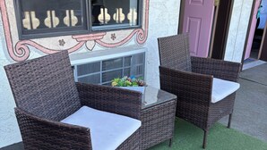A wicker outdoor seating area with a small table and cushions, set on artificial grass, in front of a building with a decorative wall and a door. - Red Bell Inn (Leavenworth)