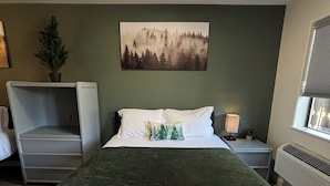 A neatly made bed with white linens and a green throw, a gray armchair, a nightstand with a lamp and a small plant, and a painting of a forest on the wall. - Red Bell Inn (Leavenworth)