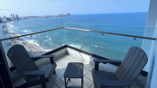 A balcony with two wooden chairs and a small table, overlooking a beach and the ocean. - The Pearl of the Pacific @ Altomare Condos (Mazatlán)