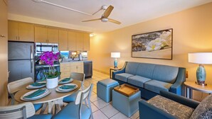 A modern kitchen with wooden cabinets, a dining area with blue chairs and a table set for two, and a living area with a blue sofa and a coffee table. - Kuleana Club - 721 (Lahaina)
