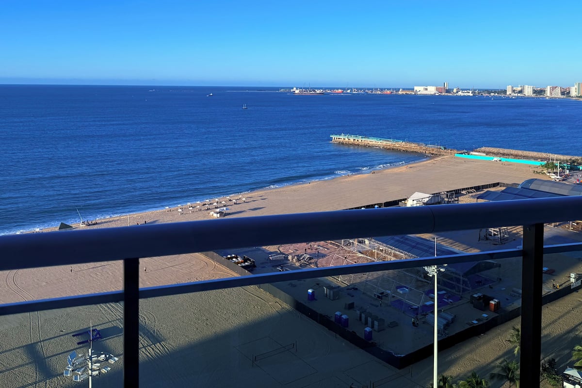 A coastal view from a balcony with a clear sky, a beach, and a distant cityscape.
