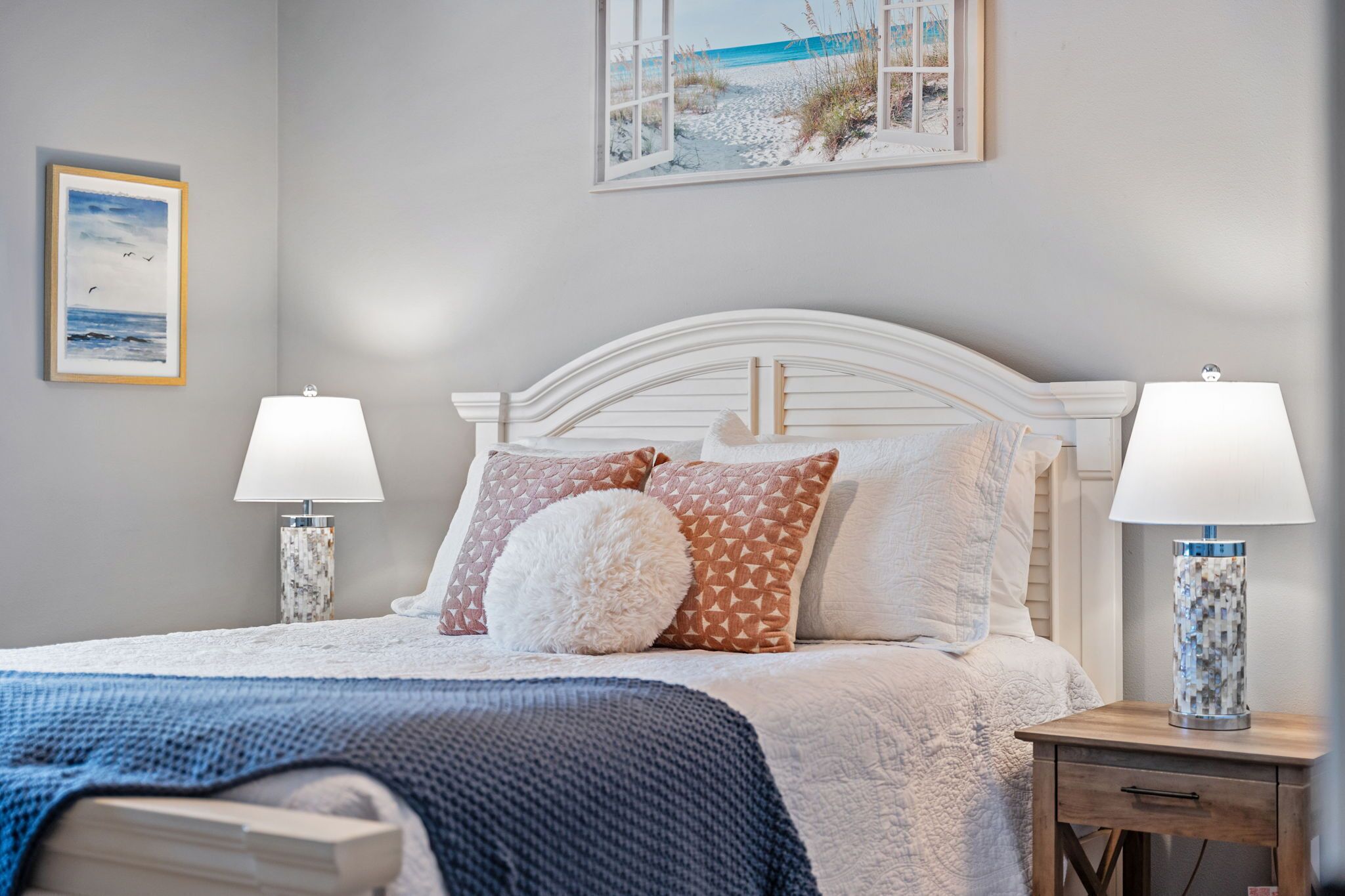 A neatly made bed with a white headboard, two bedside lamps, and a framed beach scene on the wall.