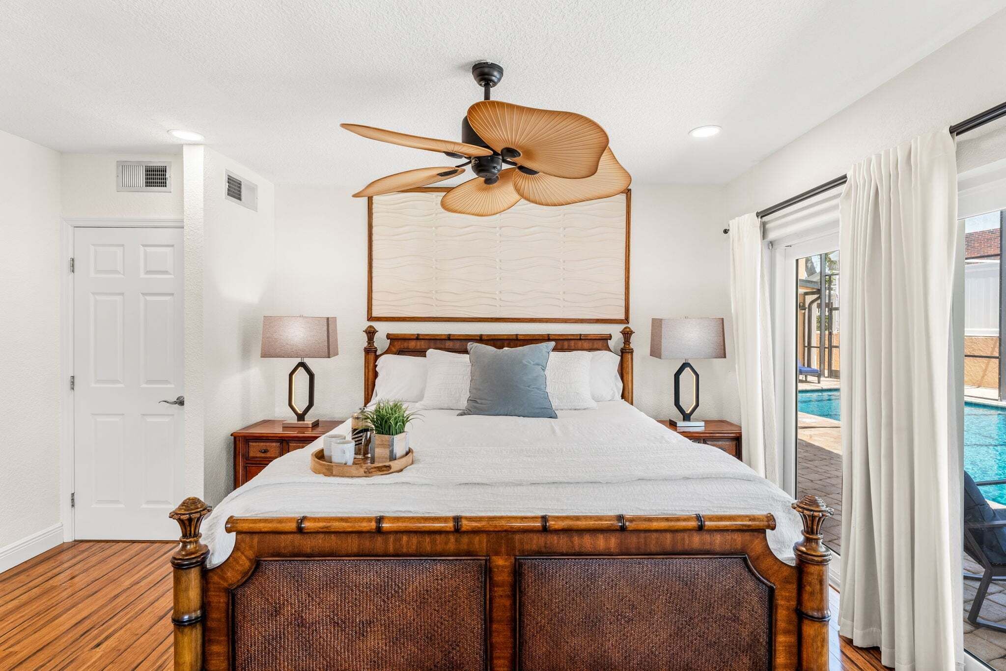 A bedroom with a wooden bed, ceiling fan, bedside lamps, and a view of an outdoor swimming pool.