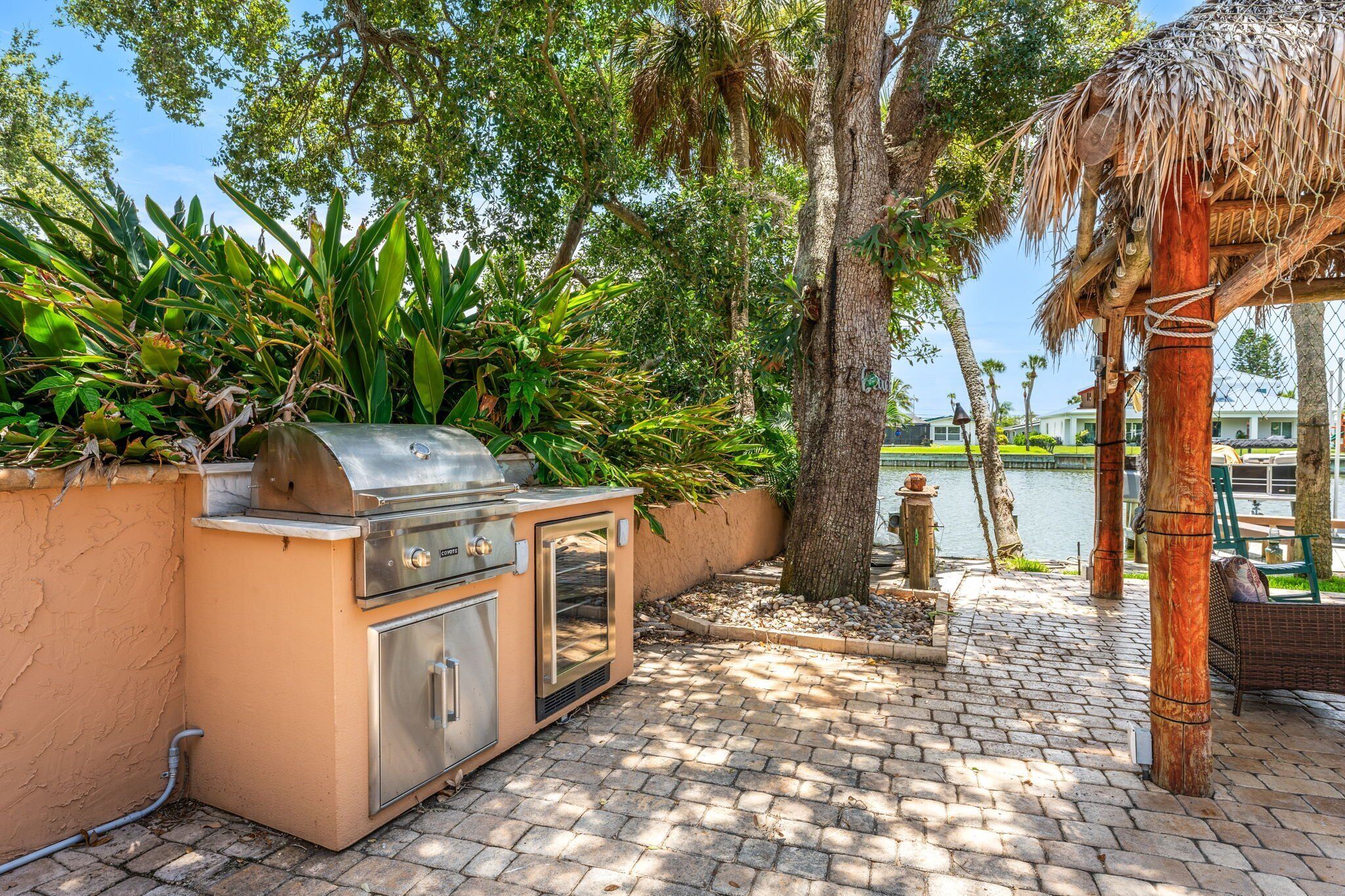 An outdoor grill area with a stone-tiled patio, surrounded by lush greenery and a view of a body of water.