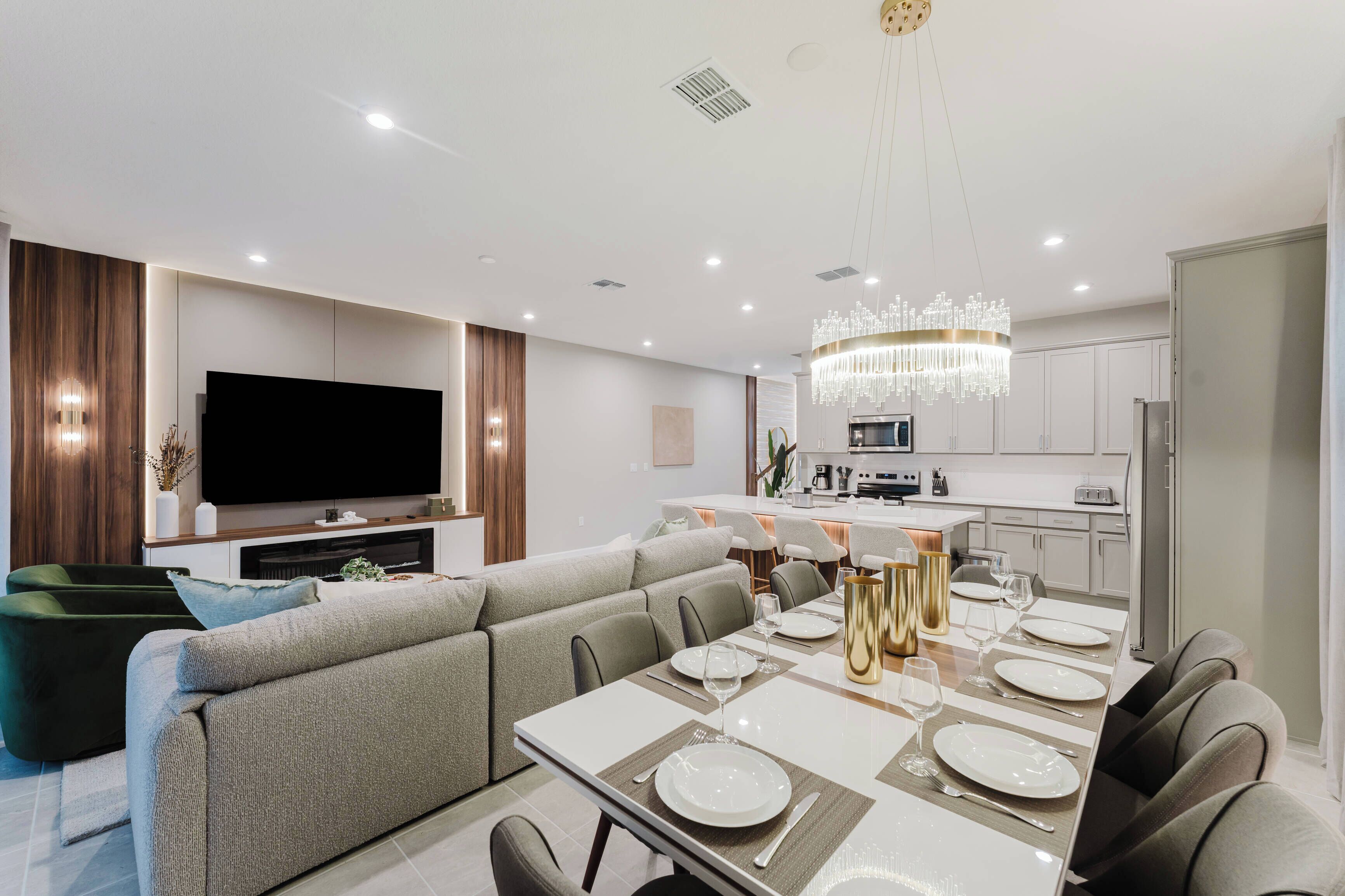 A modern living room with a large flat-screen TV, a dining area with a chandelier, and a kitchen in the background.