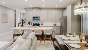 A modern kitchen with white cabinets, a central island with bar stools, and a dining area with a chandelier.