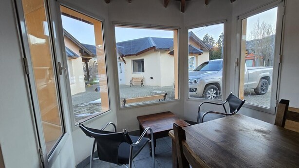 A room with a wooden table and chairs, large windows, and a view of a building and parked cars outside. - Beautiful Large Cabin – Perfect Location & Stunning Views (El Chalten)