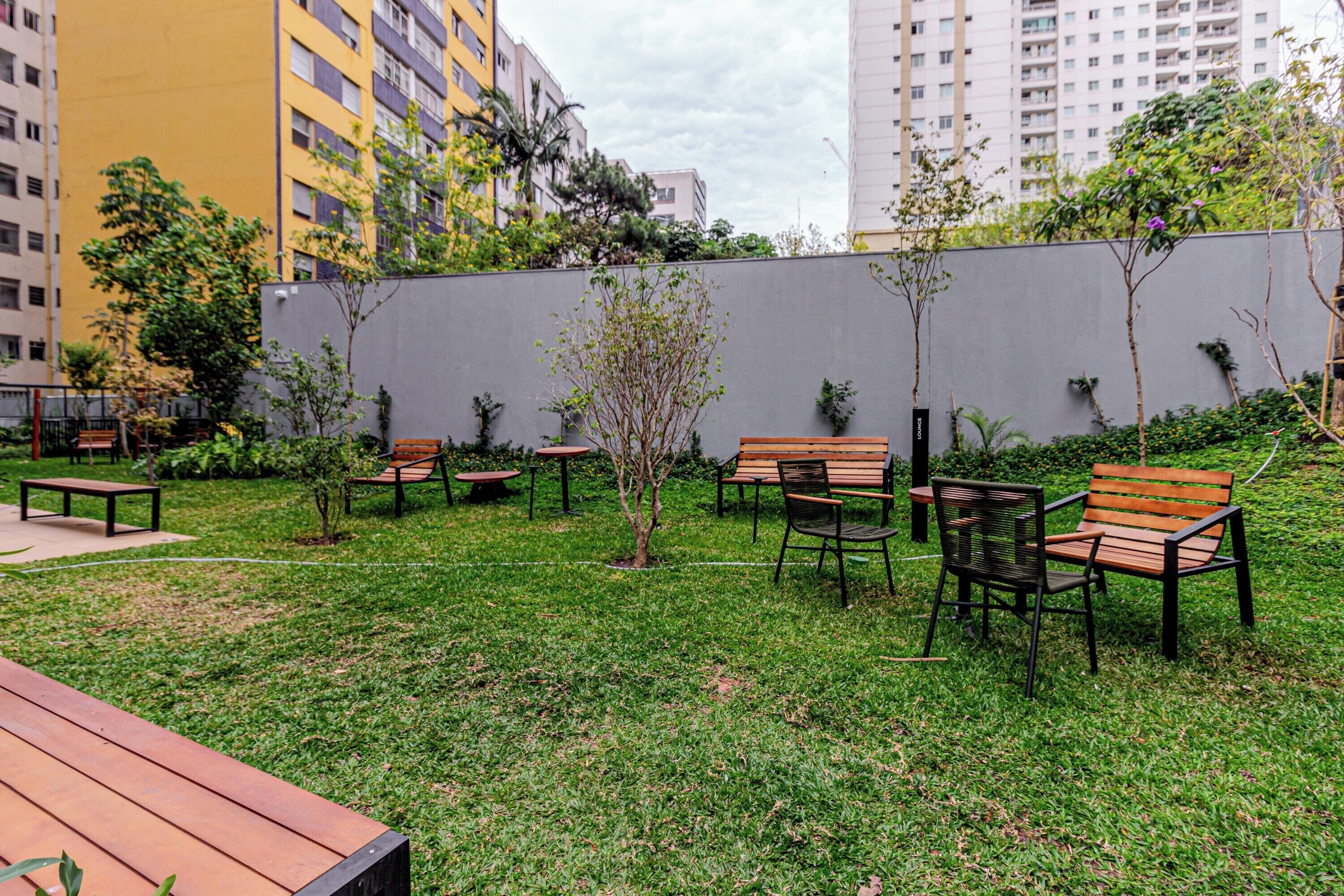 An outdoor seating area with wooden benches and tables, surrounded by a grassy lawn and small trees.