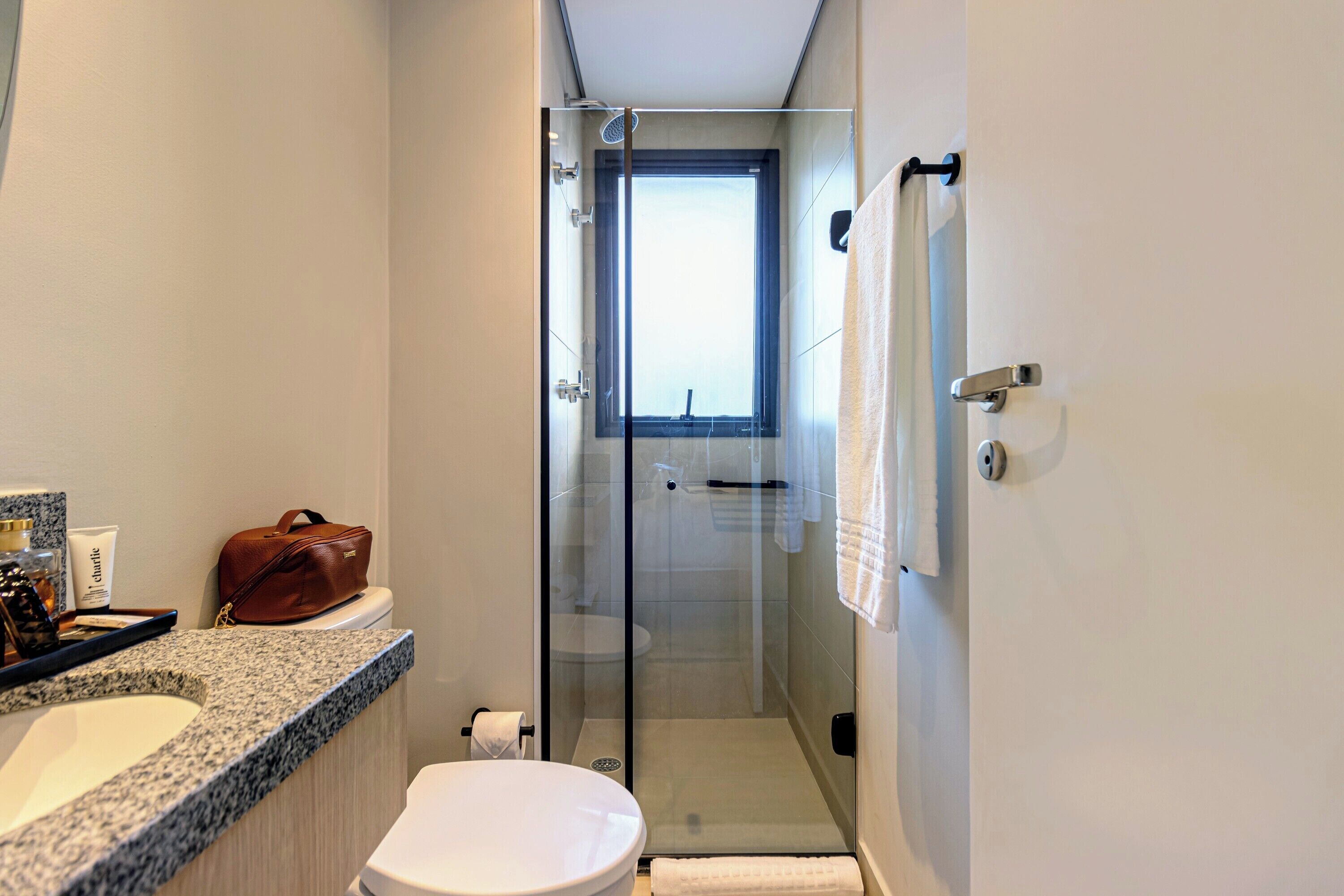 A modern bathroom with a granite countertop, a white toilet, a glass-enclosed shower, and a window providing natural light.