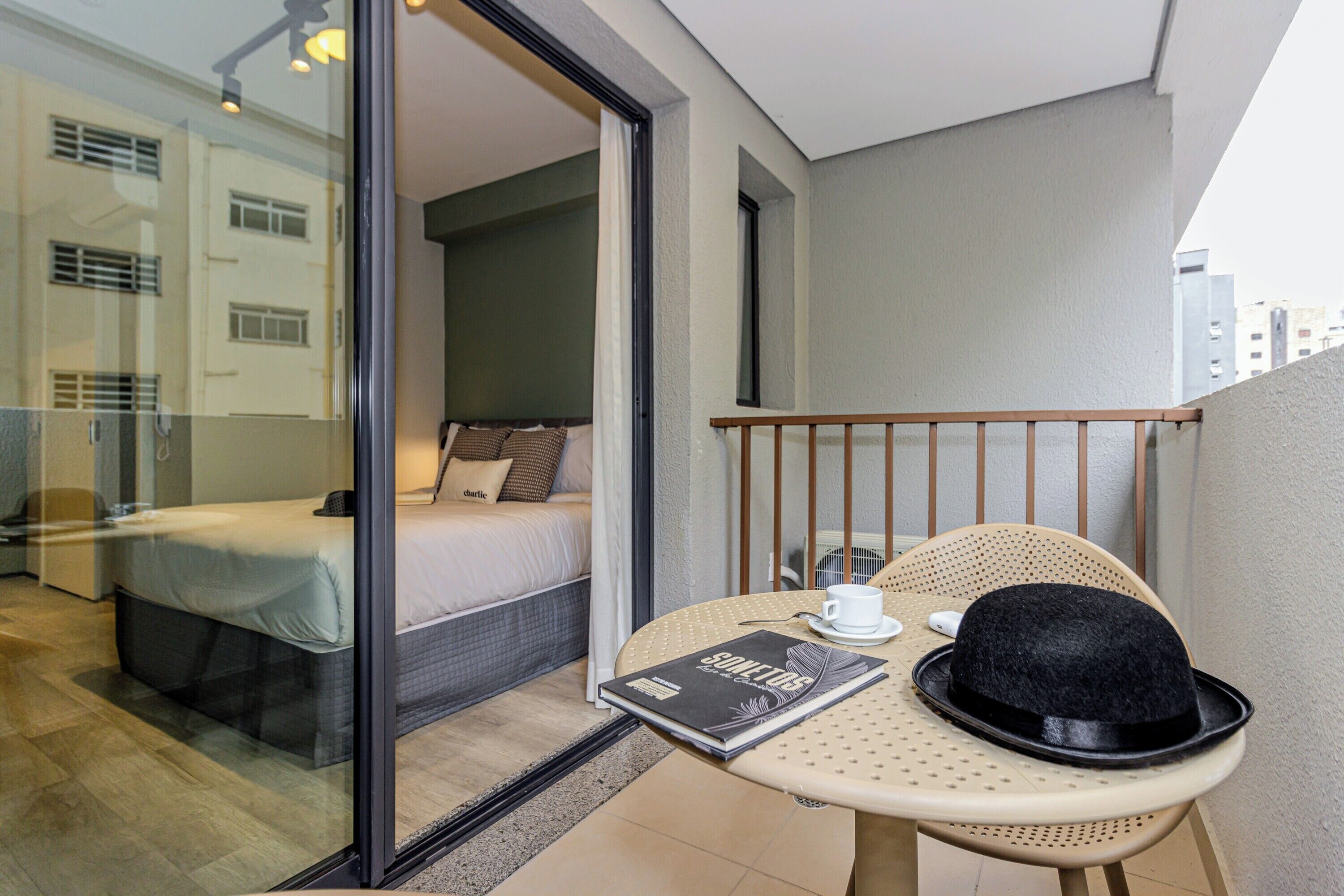 Un balcón con una cama, una mesa con un sombrero y un libro, y vistas a edificios.